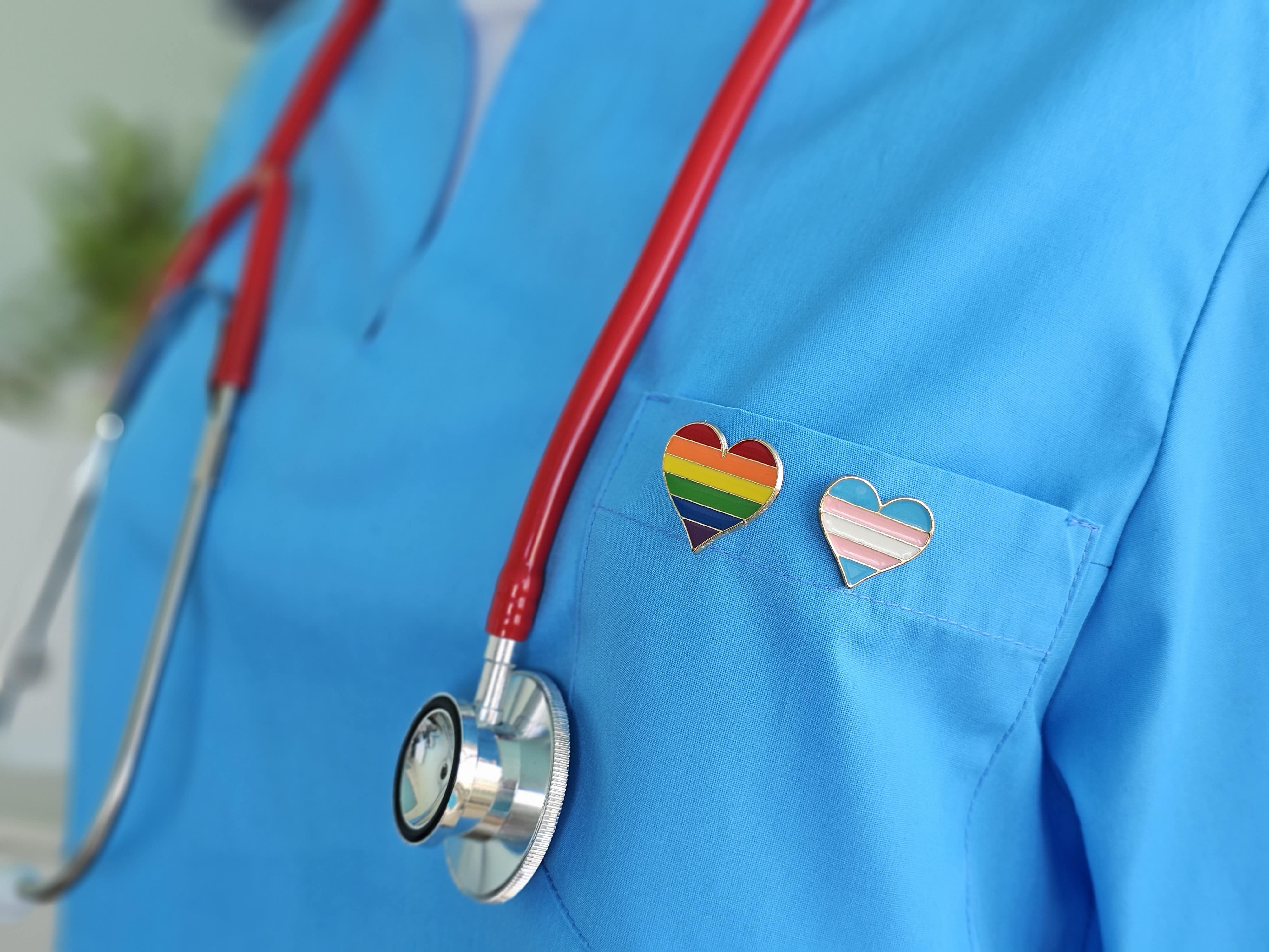 Healthcare worker wearing blue scrubs with a rainbow heart pin and a red stethoscope.