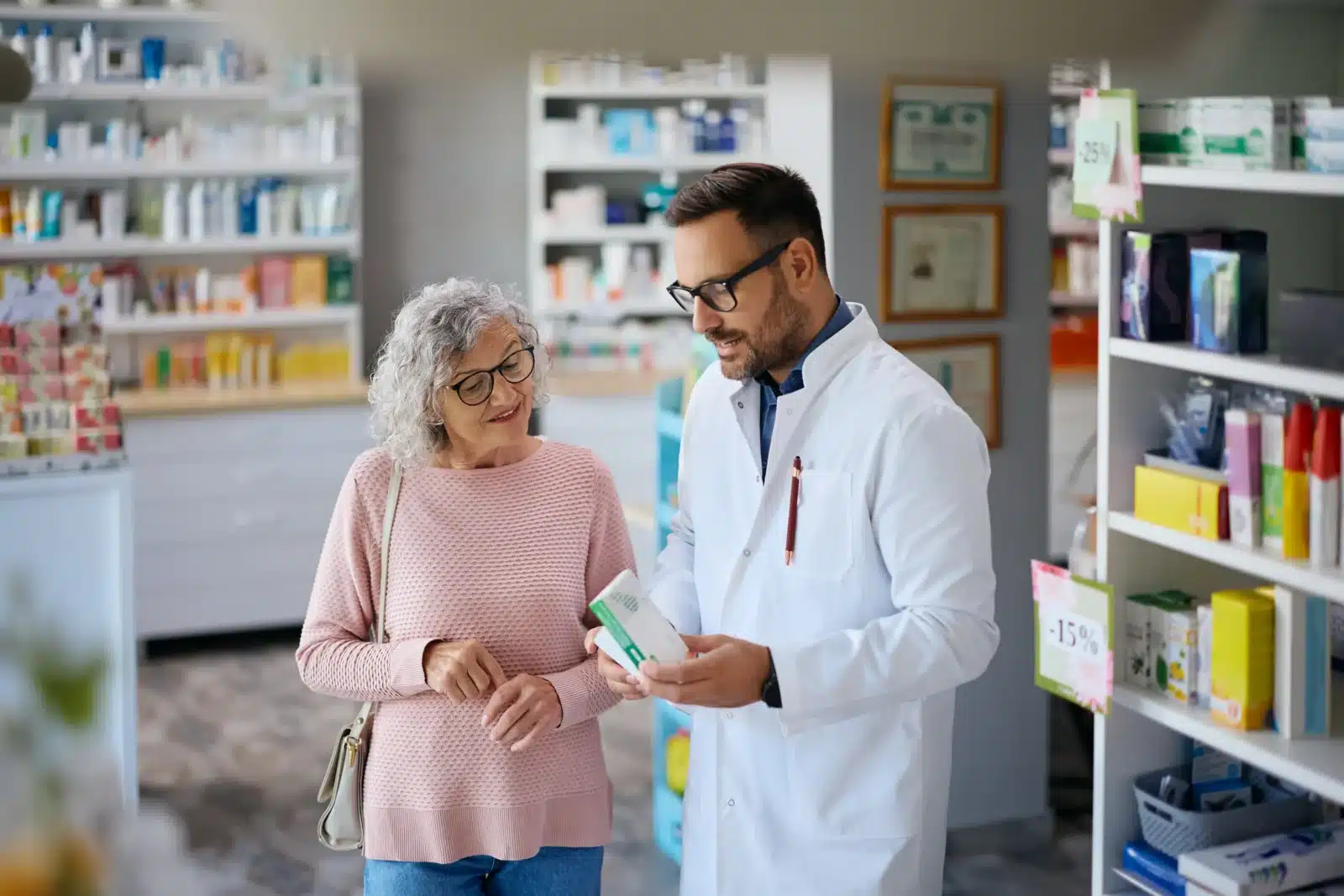 Pharmacist in a white coat advising an older woman holding a handbag inside a pharmacy.